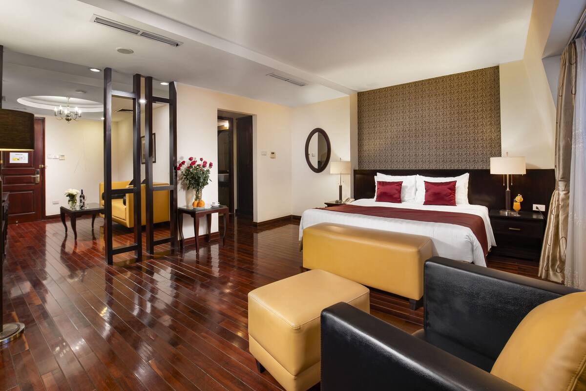 A hotel room with a large bed, a sitting area with a sofa and ottoman, a small table with a vase of flowers, and a mirror on the wall.