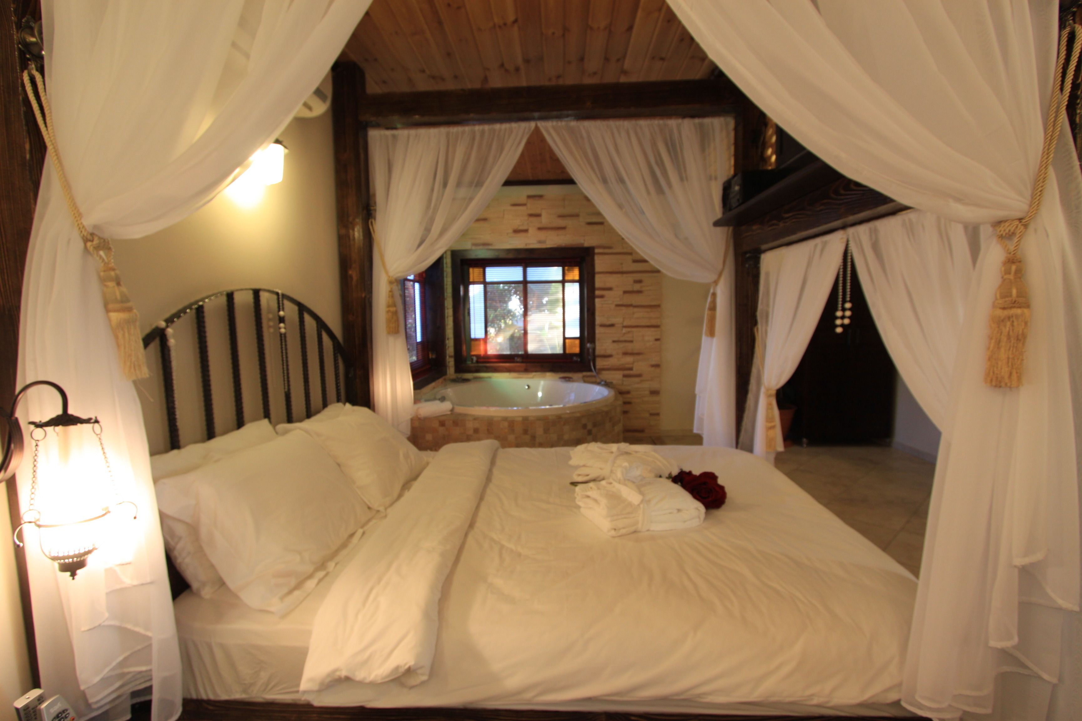 Een slaapkamer met een bed, witte beddengoed, een nachtkastje met een lamp en uitzicht op een hottub door een raam.