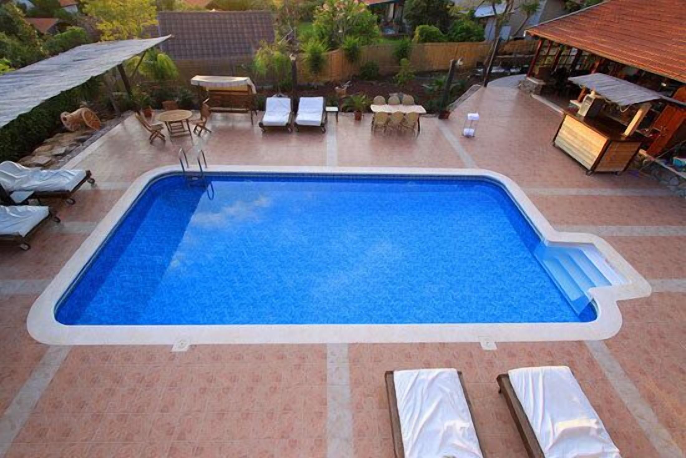 En rektangulär swimmingpool med ett kaklat poolområde, omgiven av solstolar och en möblerad, överbyggd sittplats.