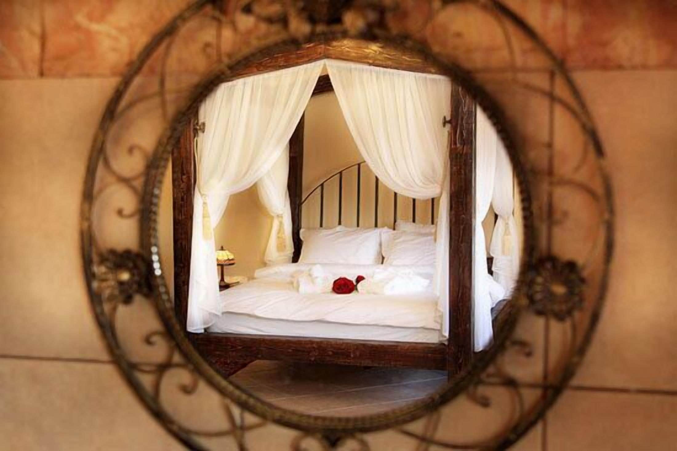 A bed with white linens and a red rose, framed by an ornate mirror with draped curtains.