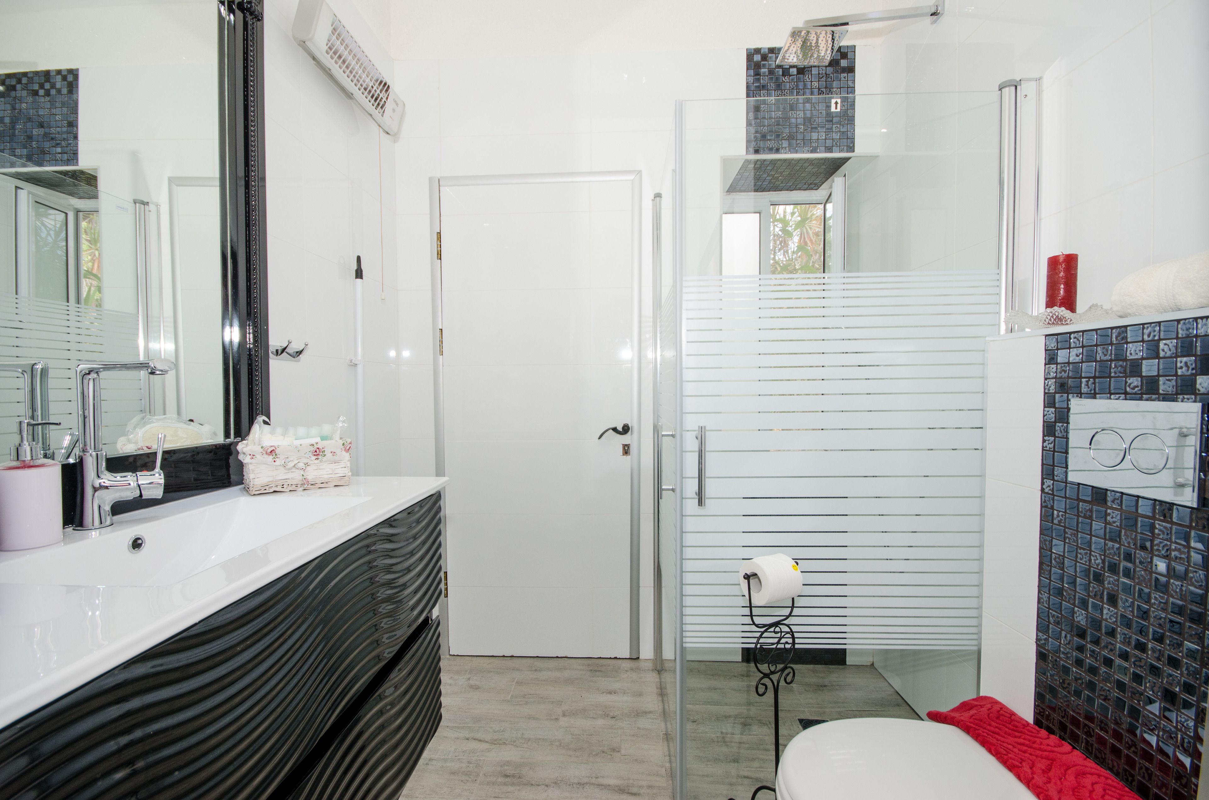 Een badkamer met een wit wastafel, spiegel en handdoekhouder. Er is een douchegedeelte met een glazen deur en een toilet met een rode handdoek.