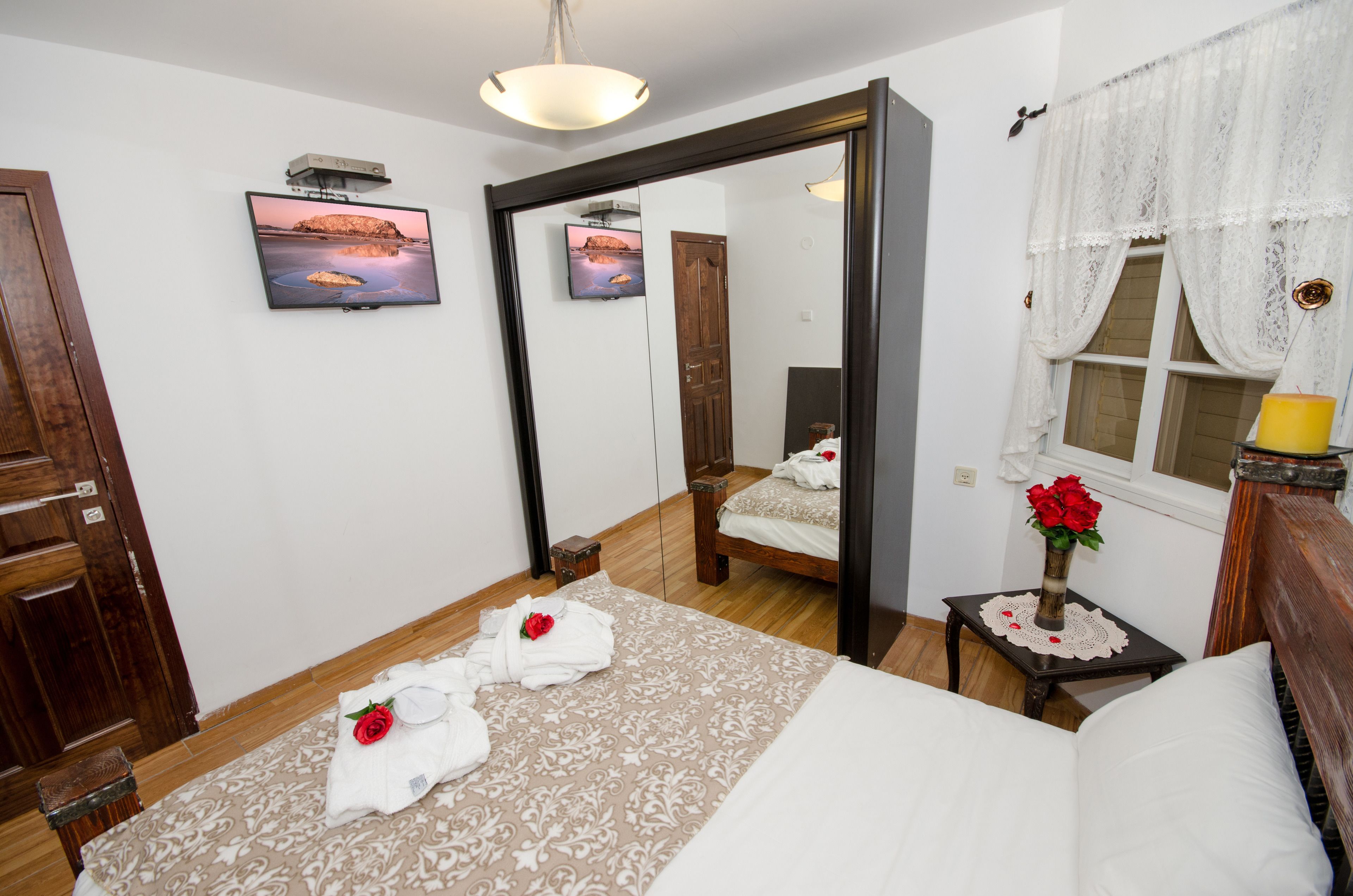 Ein Hotelzimmer mit einem großen Spiegel, einem Bett mit weißer Bettwäsche, einem Nachttisch mit einer Vase roter Rosen und einem Sonnenuntergangsbild an der Wand.