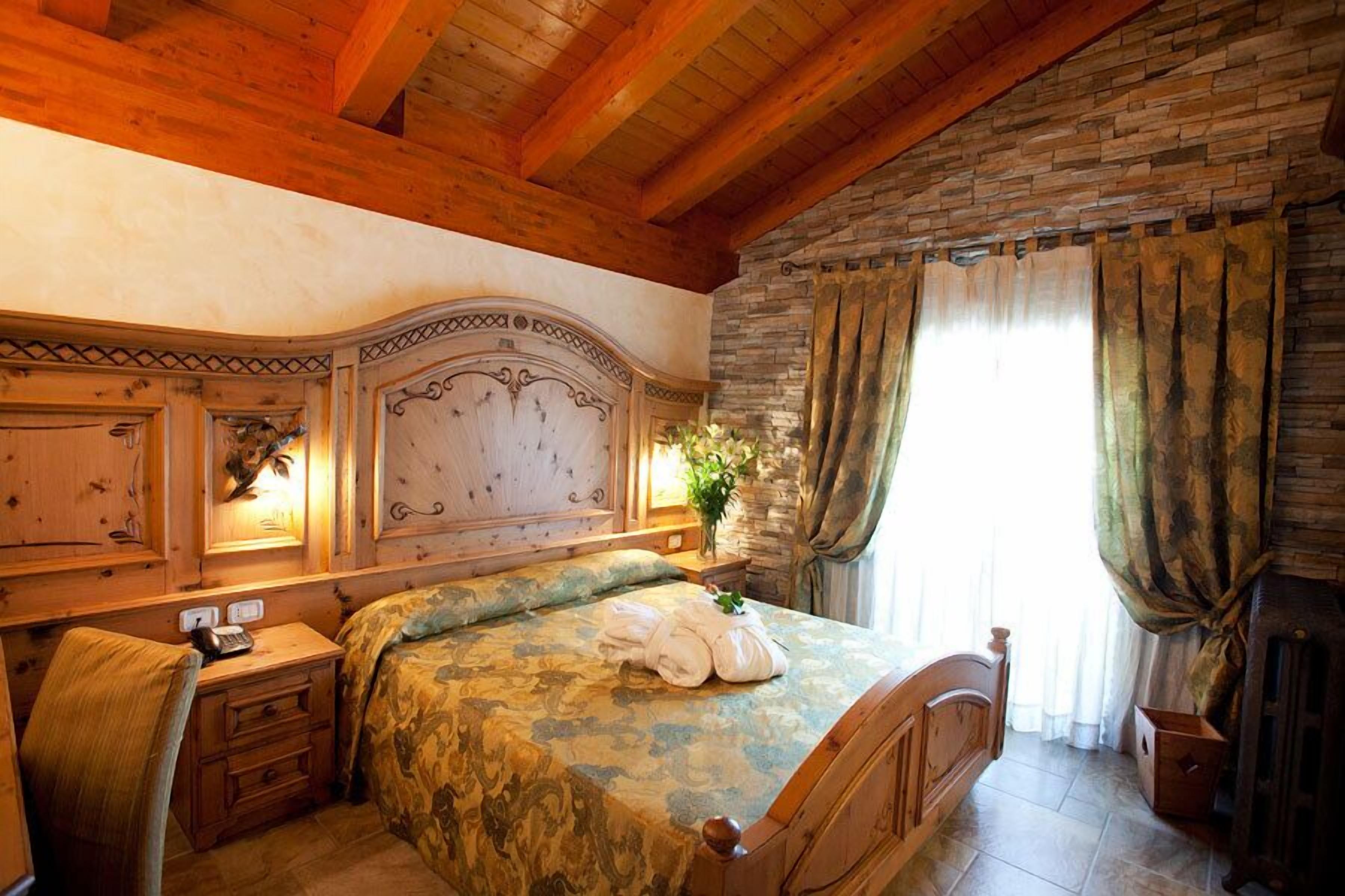 A bedroom with a wooden headboard, a bed with a patterned bedspread, a bedside table with a lamp, and a window with curtains.