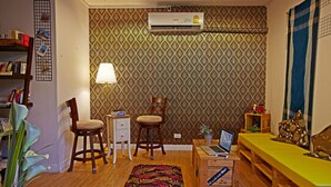 A cozy living room with a wooden floor, a patterned wallpaper, a bookshelf, a desk with a laptop, a small table with a lamp, and a sofa with cushions. - WH Hostel (Bangkok)