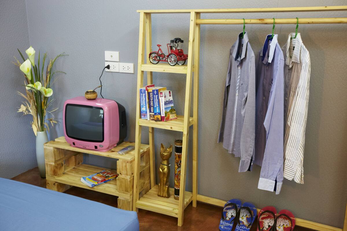 A room with a pink television on a wooden stand, a bookshelf with books and a model train, a clothing rack with striped shirts, and a pair of flip-flops on the floor.