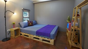 A small bedroom with a bed, a window, a floor lamp, and a wooden shelf with various items. - WH Hostel (Bangkok)