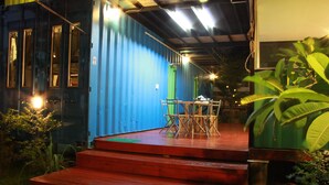 A blue container house with a wooden deck and steps leading to a table and chairs. - WH Hostel (Bangkok)