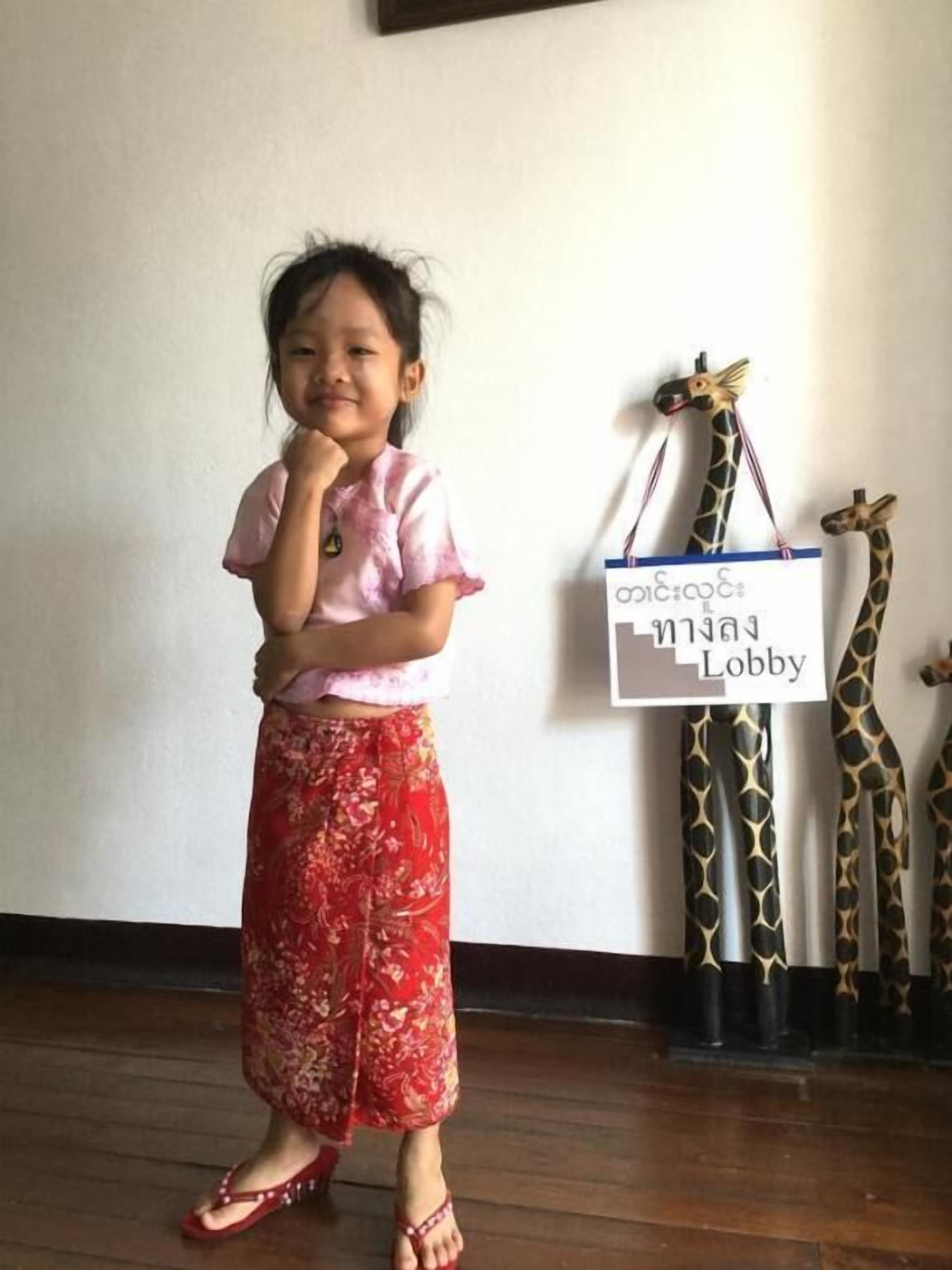 A child in a pink top and red skirt standing in a room with wooden flooring and a white wall. There is a sign on the wall that reads "Mian Lobby".