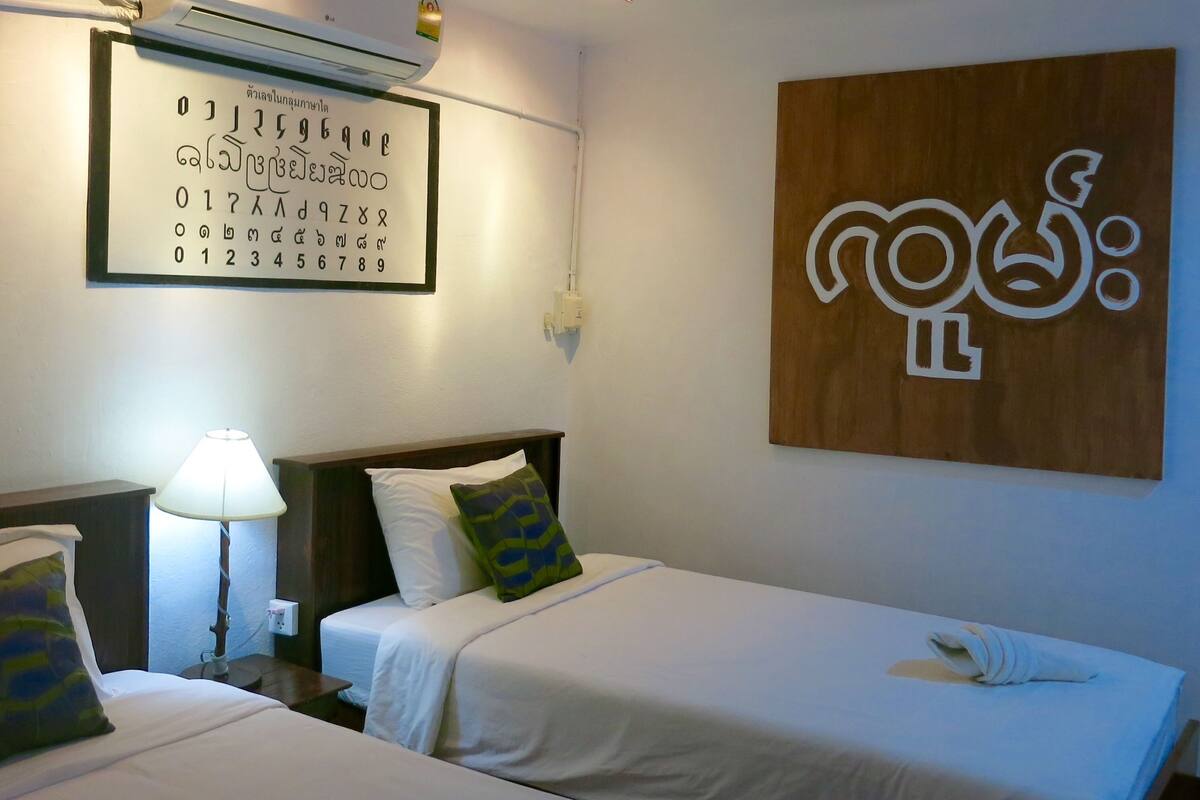 A hotel room with two beds, a wall-mounted air conditioner, a wall clock, and a wooden artwork.