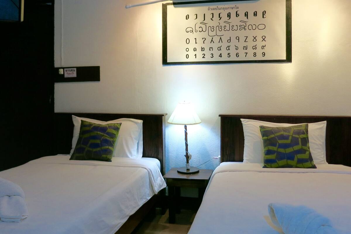 A hotel room with two beds, each with a pillow and a bedside table with a lamp. A wall clock and a framed picture are also visible.