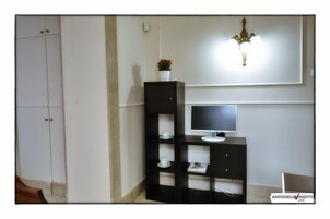 Una habitación con un televisor sobre un mueble bajo, una estantería negra con una planta y una pared blanca con una lámpara.