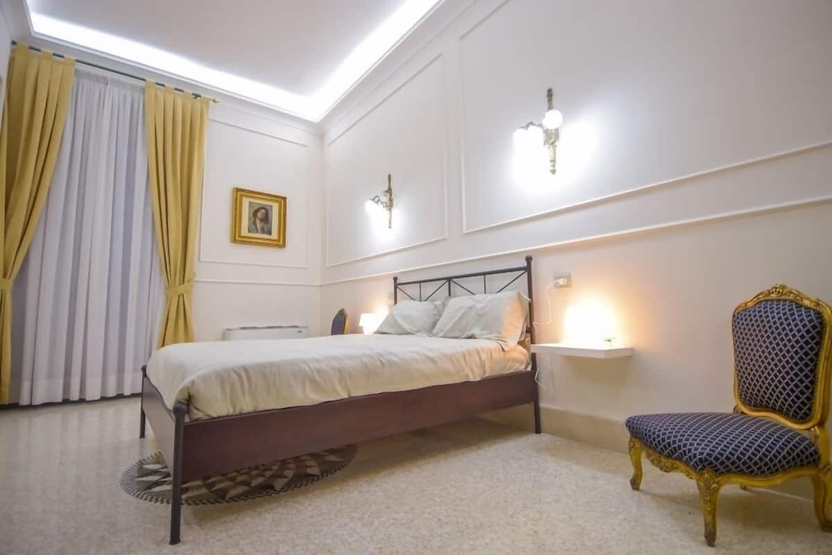 Un dormitori amb llit, cadira, tauleta de nit i un quadre a la paret.