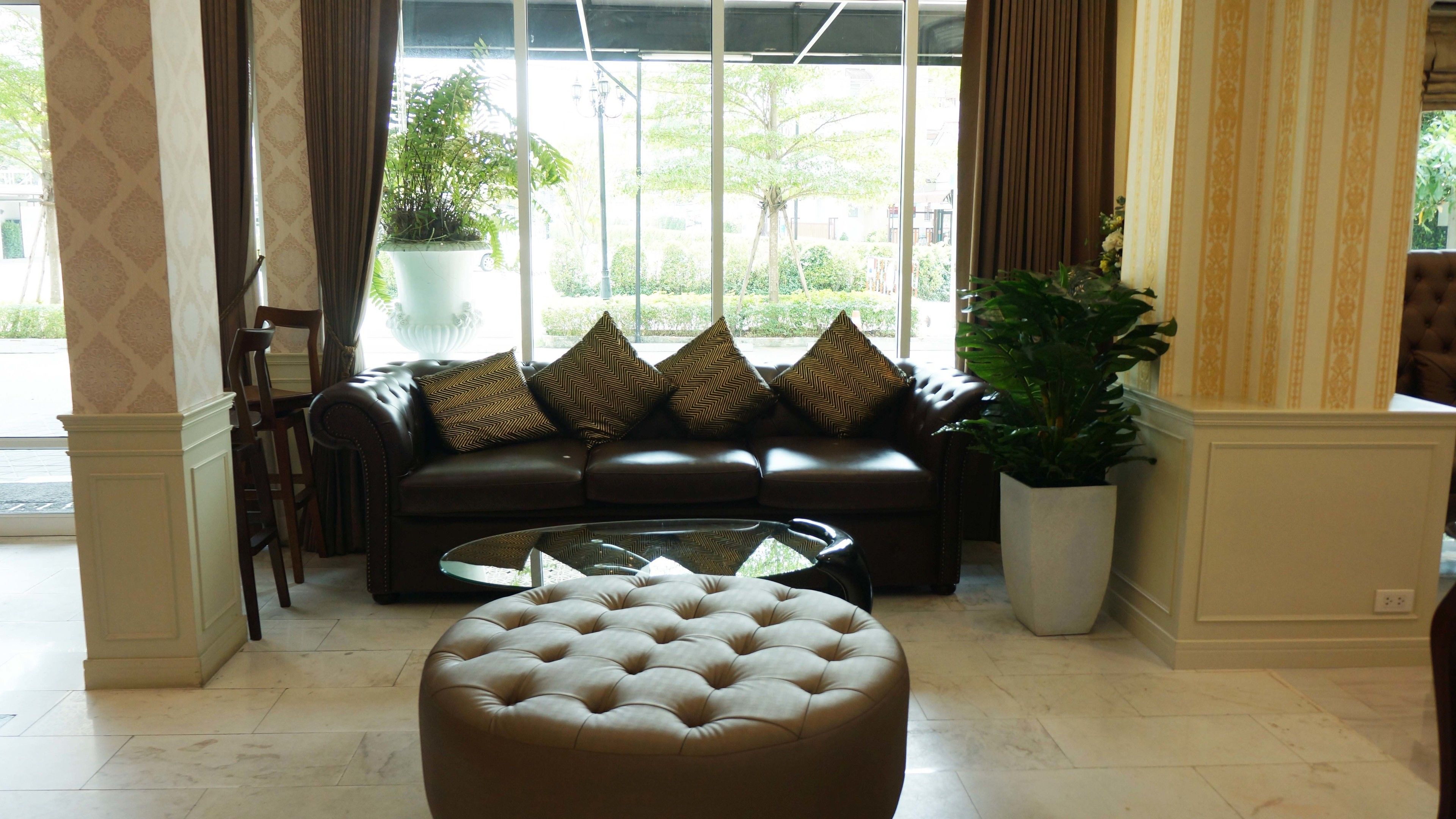 A modern living room with a leather sofa, a glass coffee table, and a tufted ottoman. There is a large window with curtains, a potted plant, and a view of the outside.