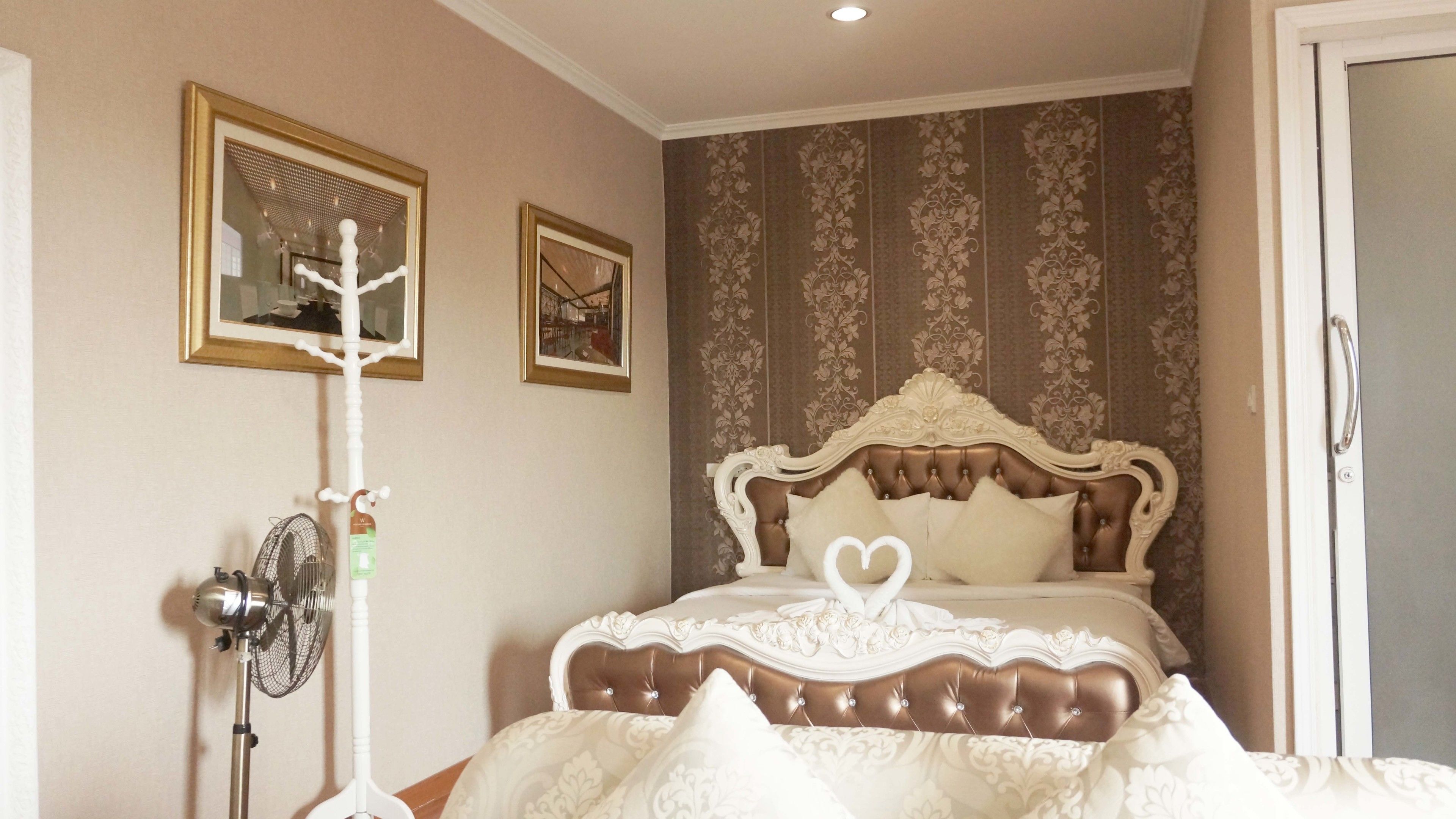A bedroom with a large bed, a standing fan, and two framed pictures on the wall.