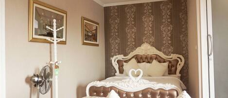 A bedroom with a large bed, a standing fan, and two framed pictures on the wall.
