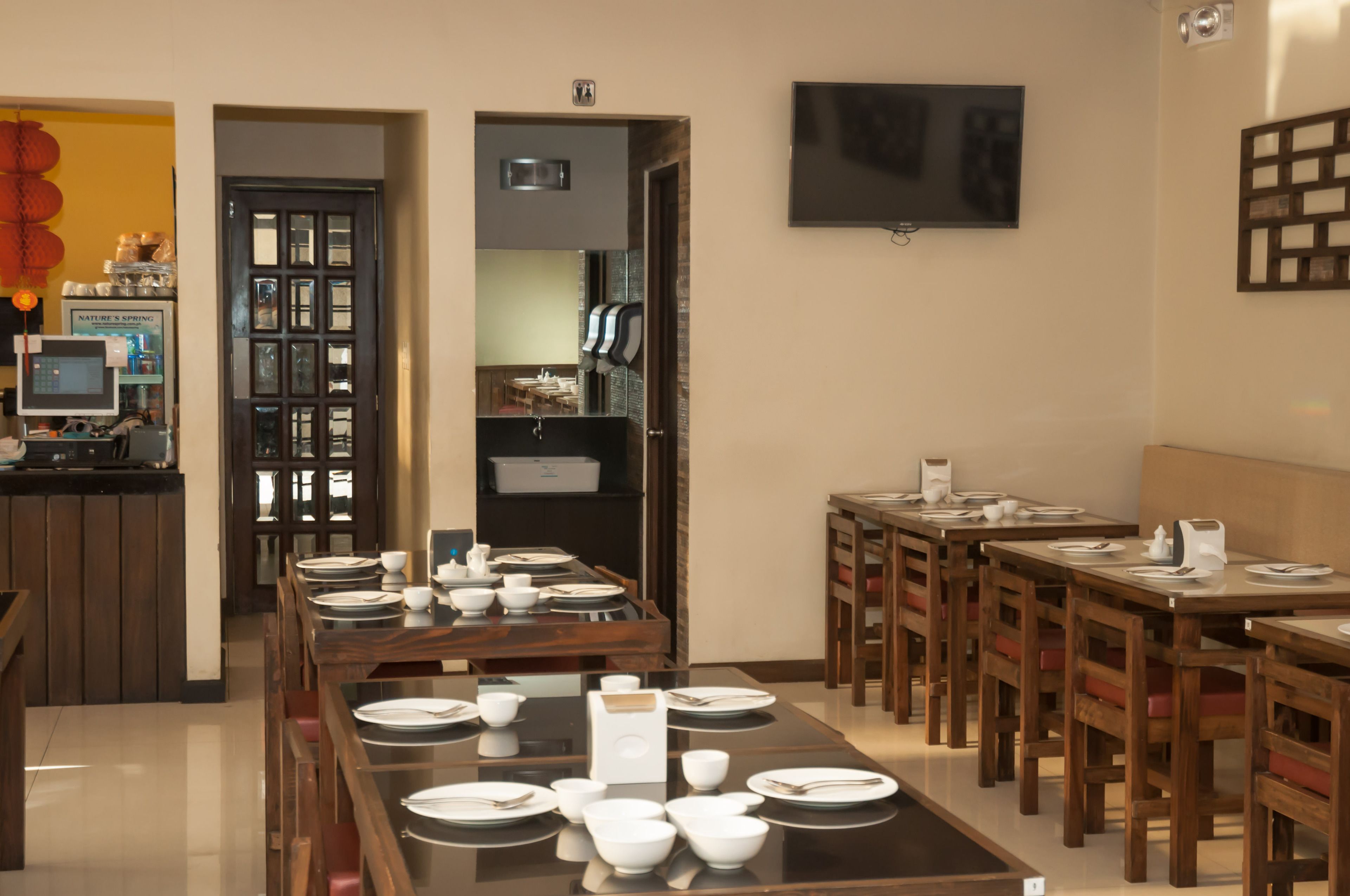 A dining area with multiple tables set for service, a television mounted on the wall, and a kitchen area visible through an open door.