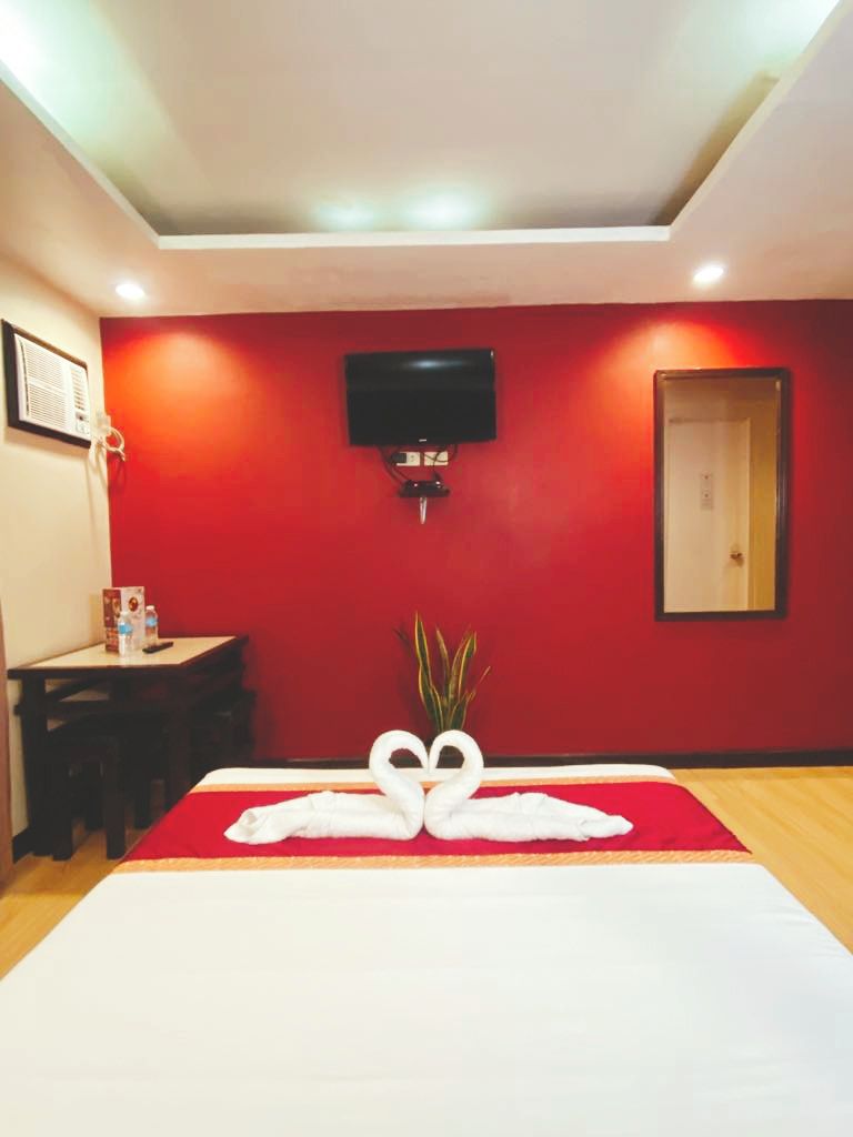 Habitación de hotel con pared roja, un televisor de pantalla plana montado en la pared, cama con colcha blanca y una mesita con una planta y una botella de agua.