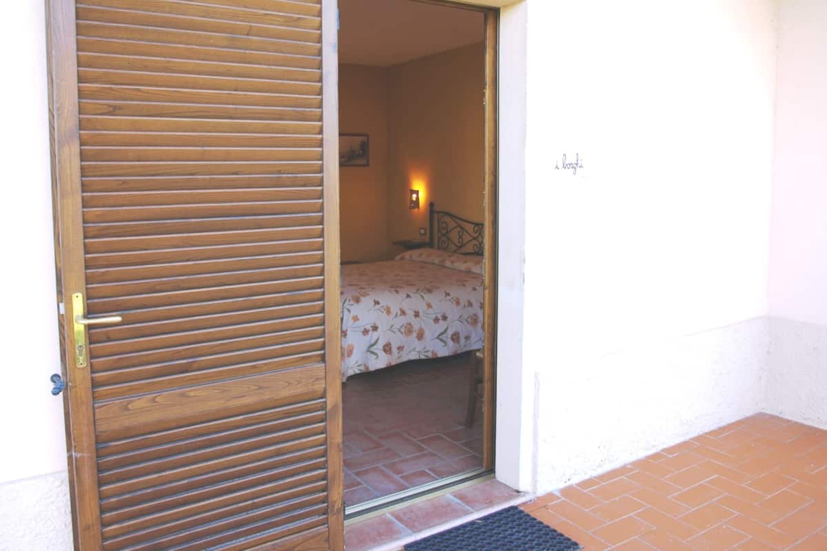 Habitació amb porta de fusta, llit amb roba de llit floral, llum penjada a la paret i una petita catifes al terra.