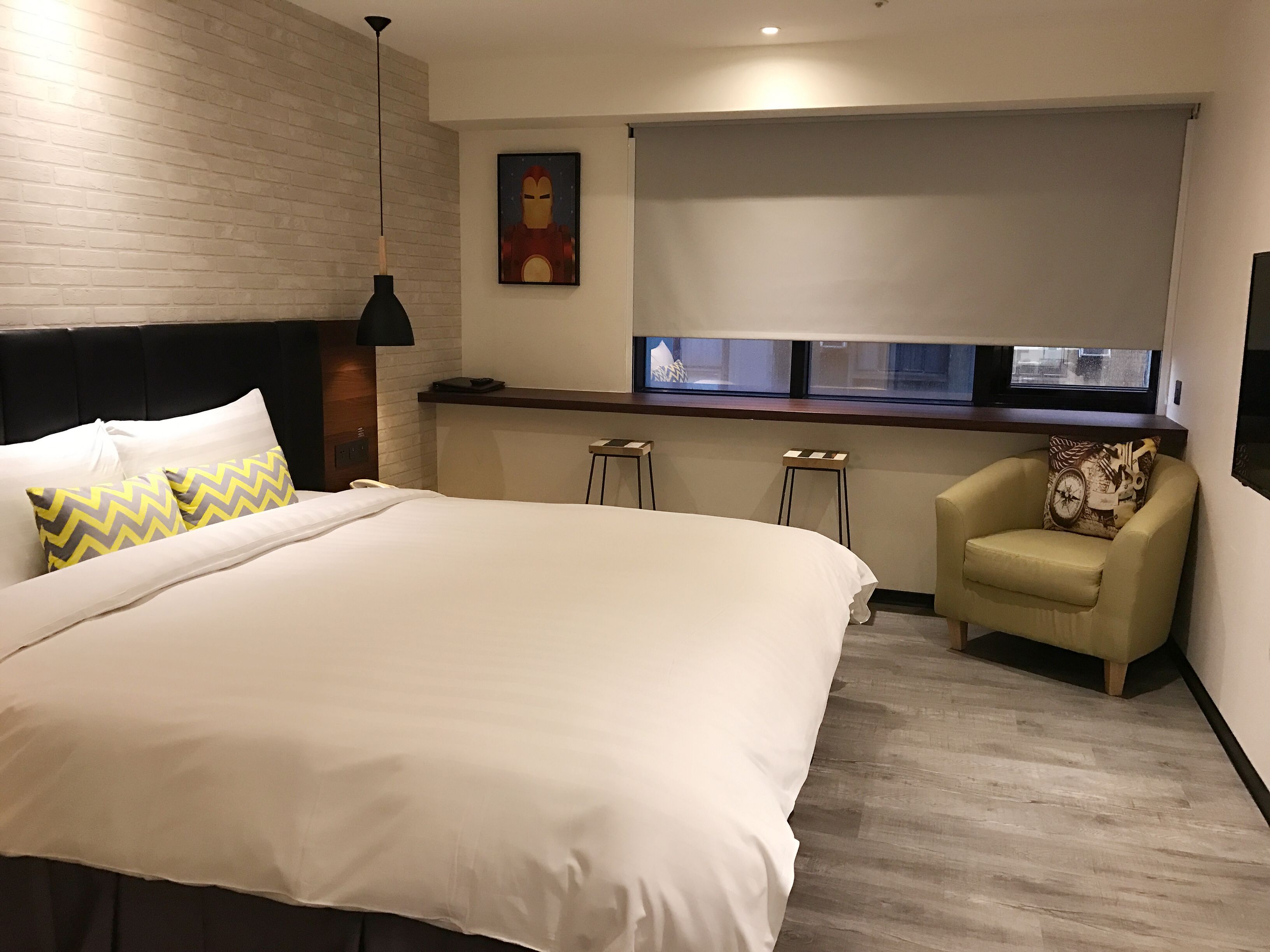 A hotel room with a large bed, a desk, two stools, a chair, a window with a view, and a wall-mounted artwork.