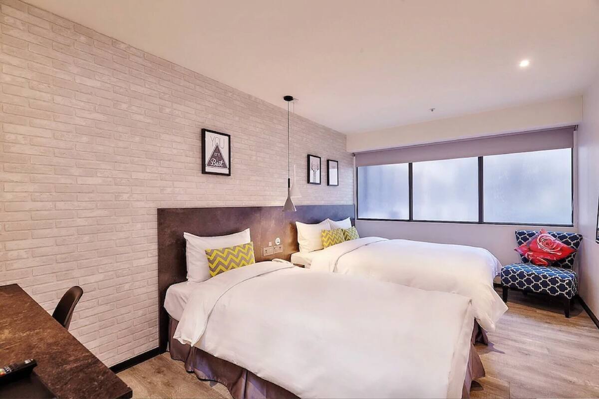 A hotel room with two beds, a brick wall, a large window, and a chair with a red cushion.