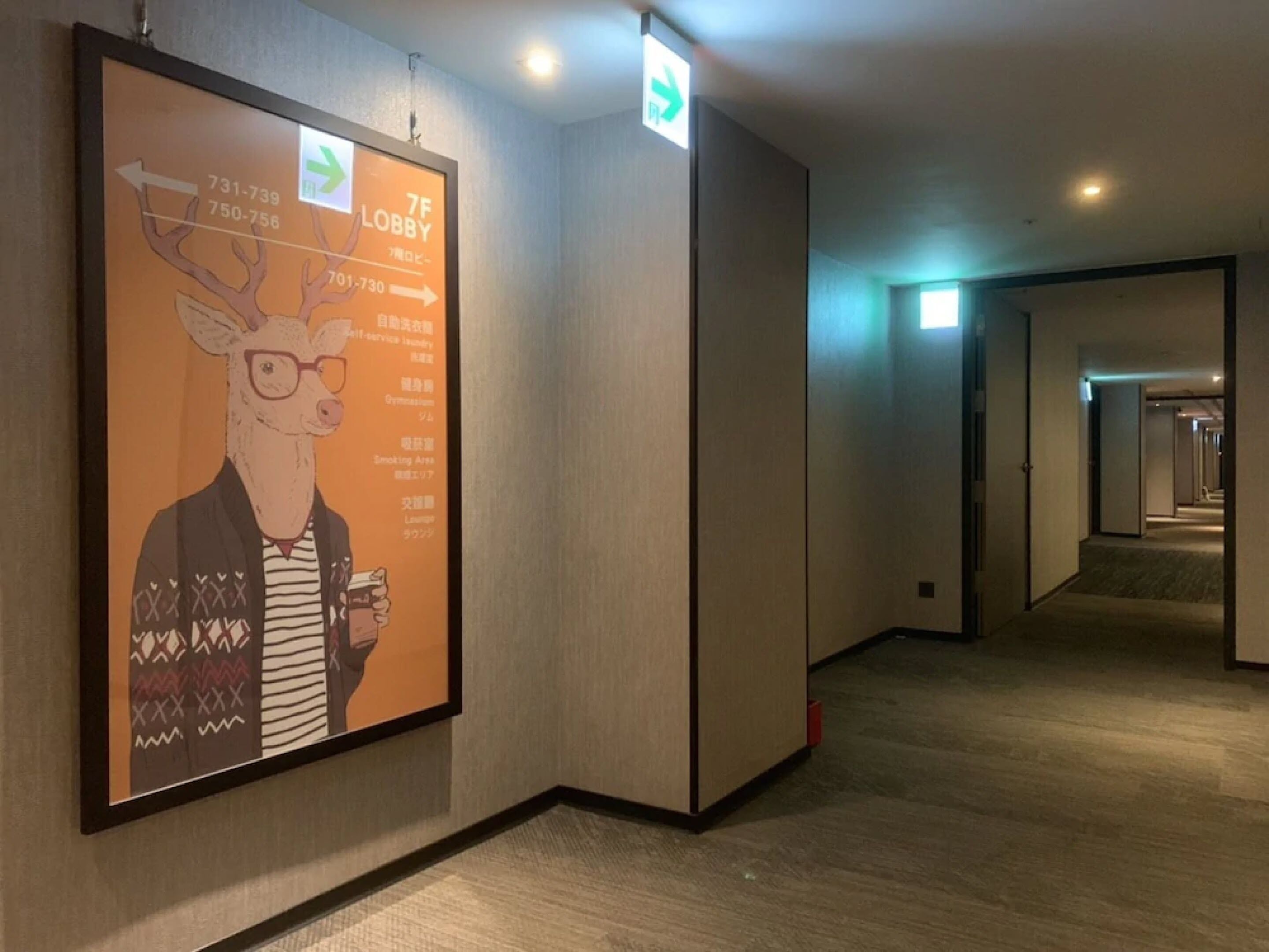 A hallway with a framed poster on the left, a door with an exit sign, and a carpeted floor.