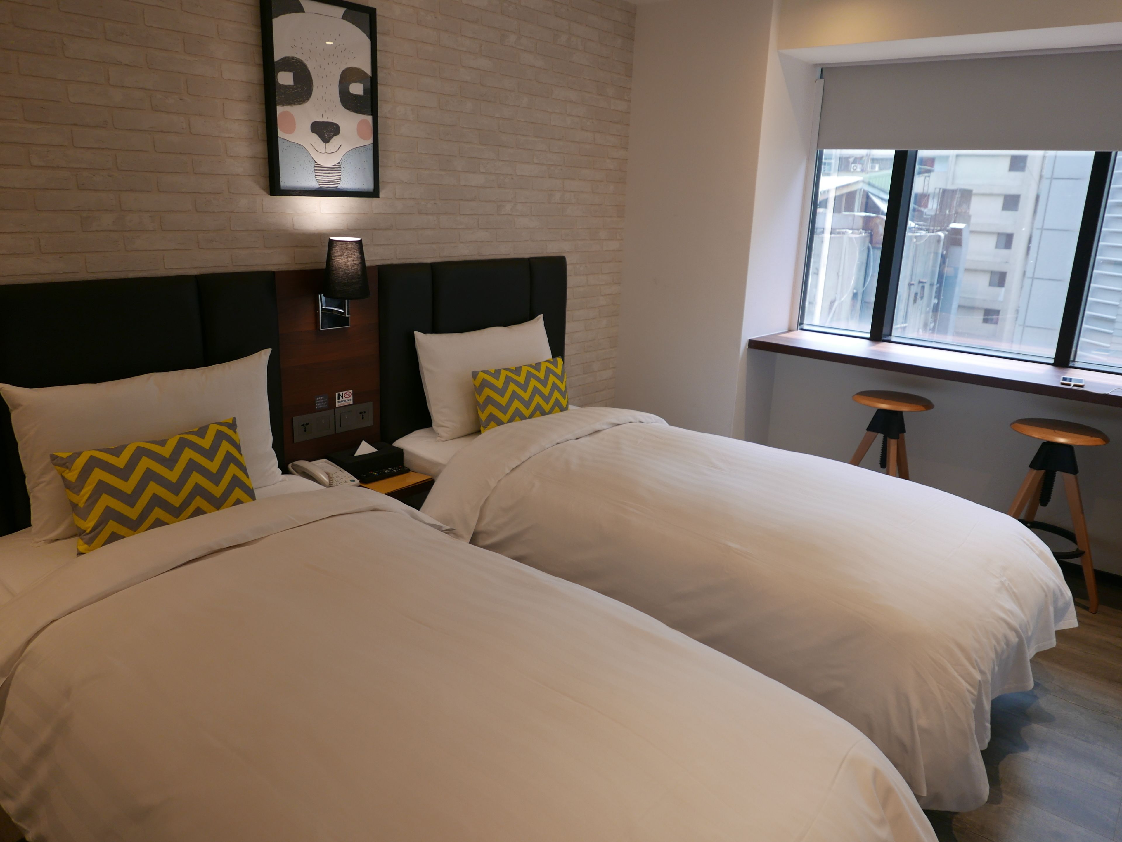 Une chambre d’hôtel avec deux lits, un mur en briques, un tableau représentant un animal, une table de chevet avec une lampe et une fenêtre donnant sur des immeubles.