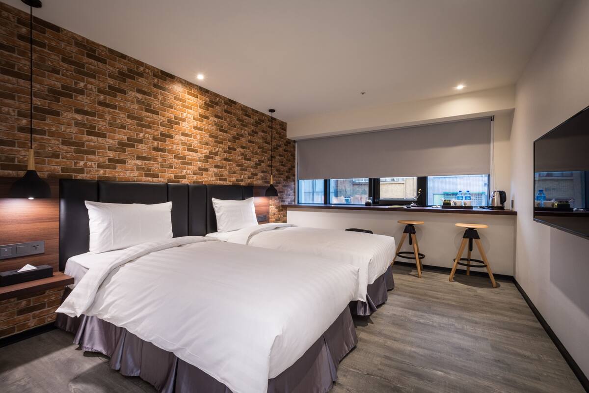 A modern hotel room with a brick accent wall, a large bed with white linens, a desk with two monitors, and a bar area with stools.