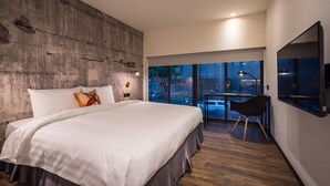 A modern bedroom with a large bed, a concrete accent wall, a desk with a chair, and a large window offering a view of the outdoors. - Goldinn Hotel (Taipei)