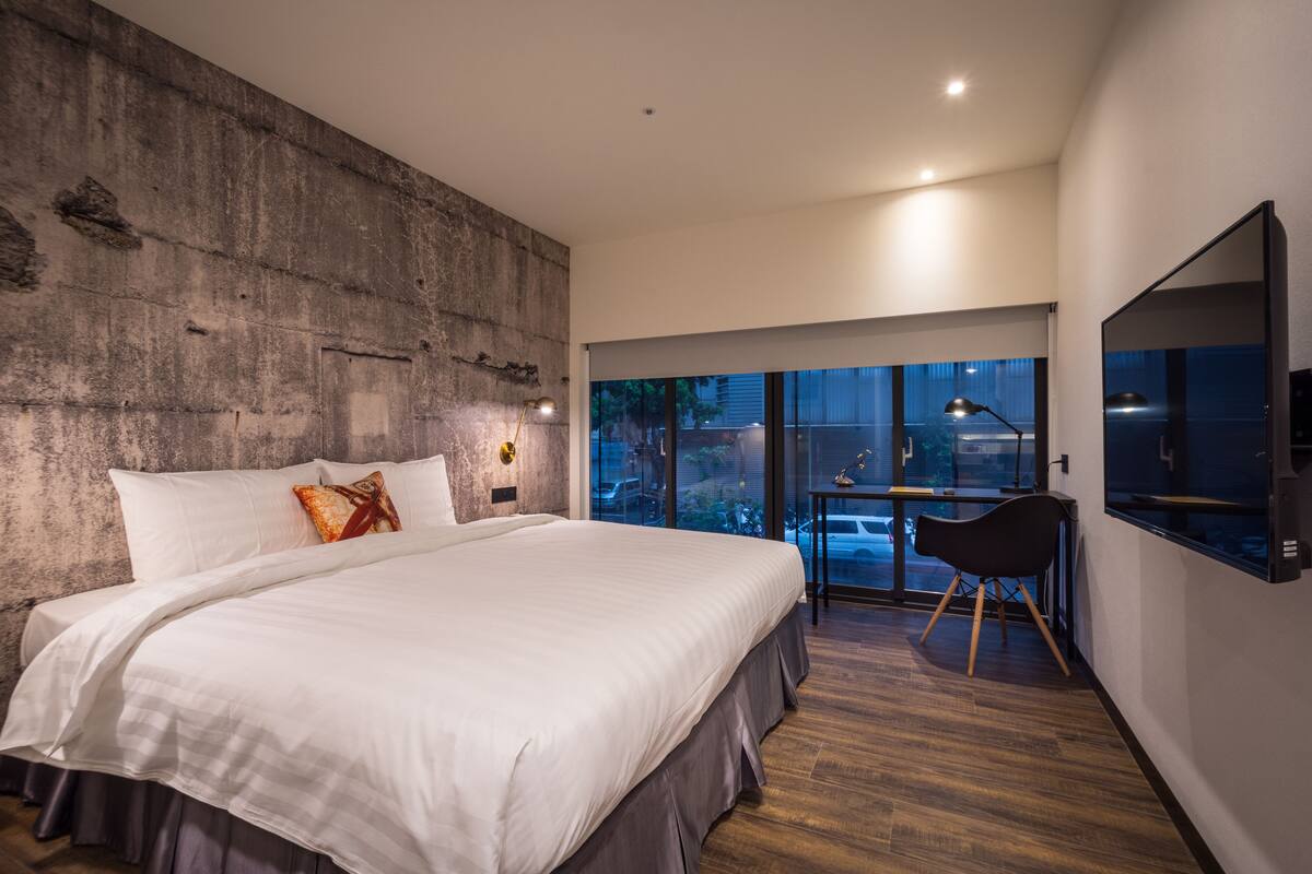 A modern bedroom with a large bed, a concrete accent wall, a desk with a chair, and a large window offering a view of the outdoors.