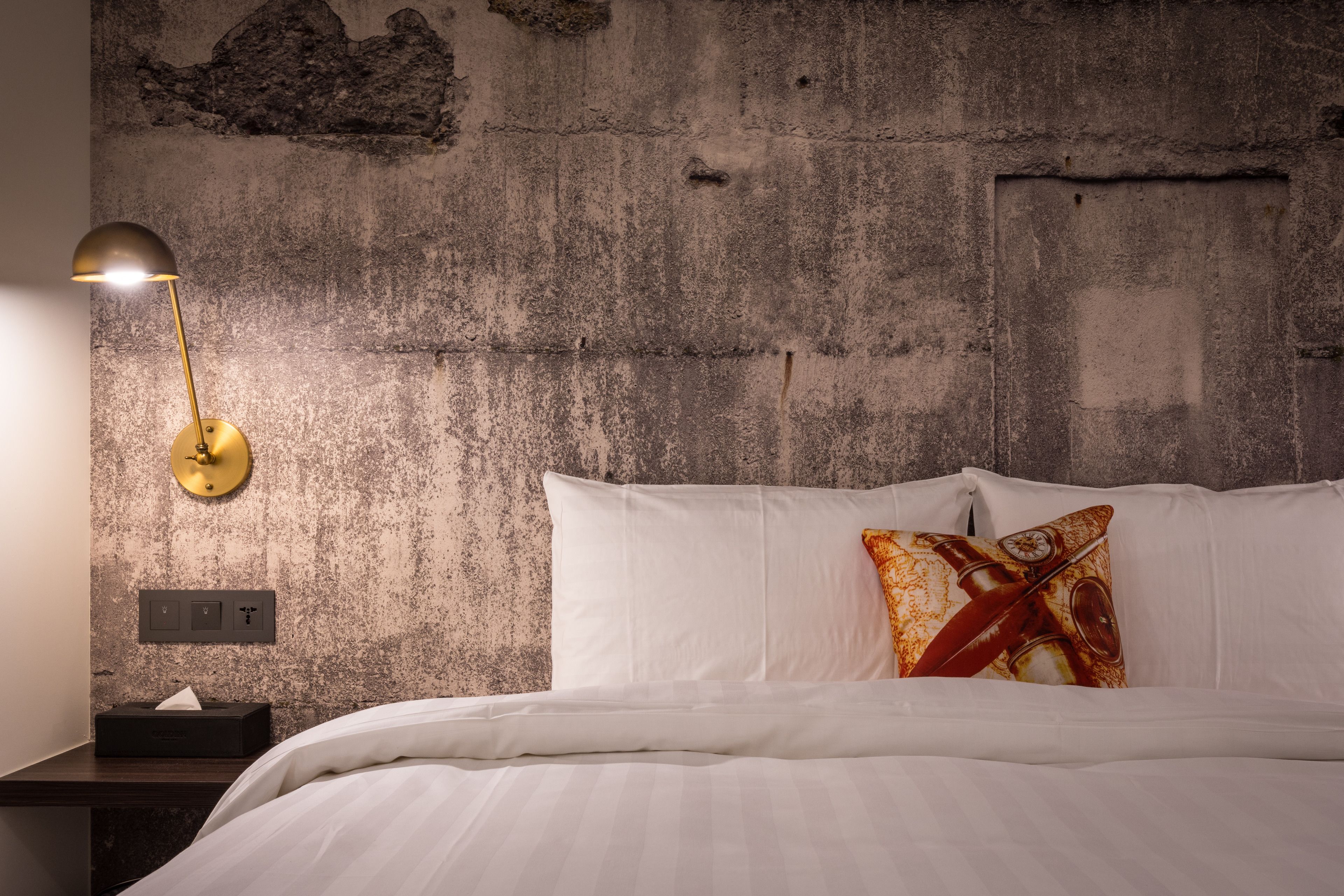 Een slaapkamer met een betonnen muur, een bedlampje, een bed met wit beddengoed en een kussen met een rood en geel dessin.