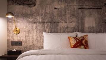 Een slaapkamer met een betonnen muur, een bedlampje, een bed met wit beddengoed en een kussen met een rood en geel dessin.