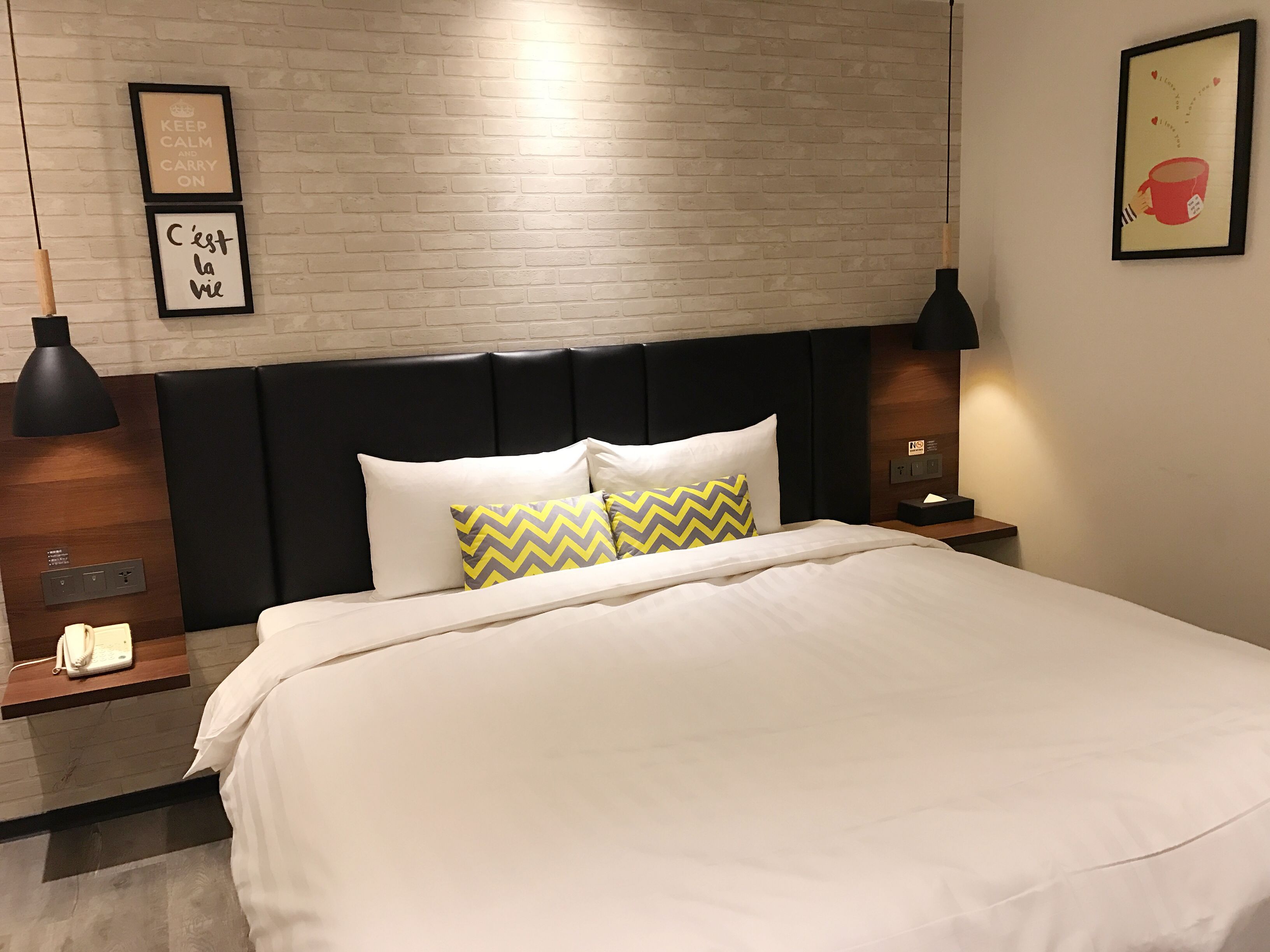 A hotel room with a brick-patterned wall, a black headboard, a white bedspread, two pillows with a chevron pattern, and two bedside lamps.