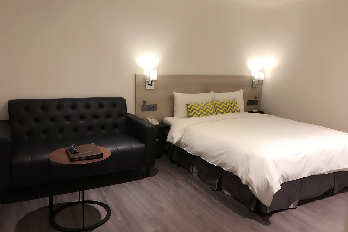 A hotel room with a black leather sofa, a small round table, a bed with white bedding and patterned pillows, and two wall-mounted lamps.