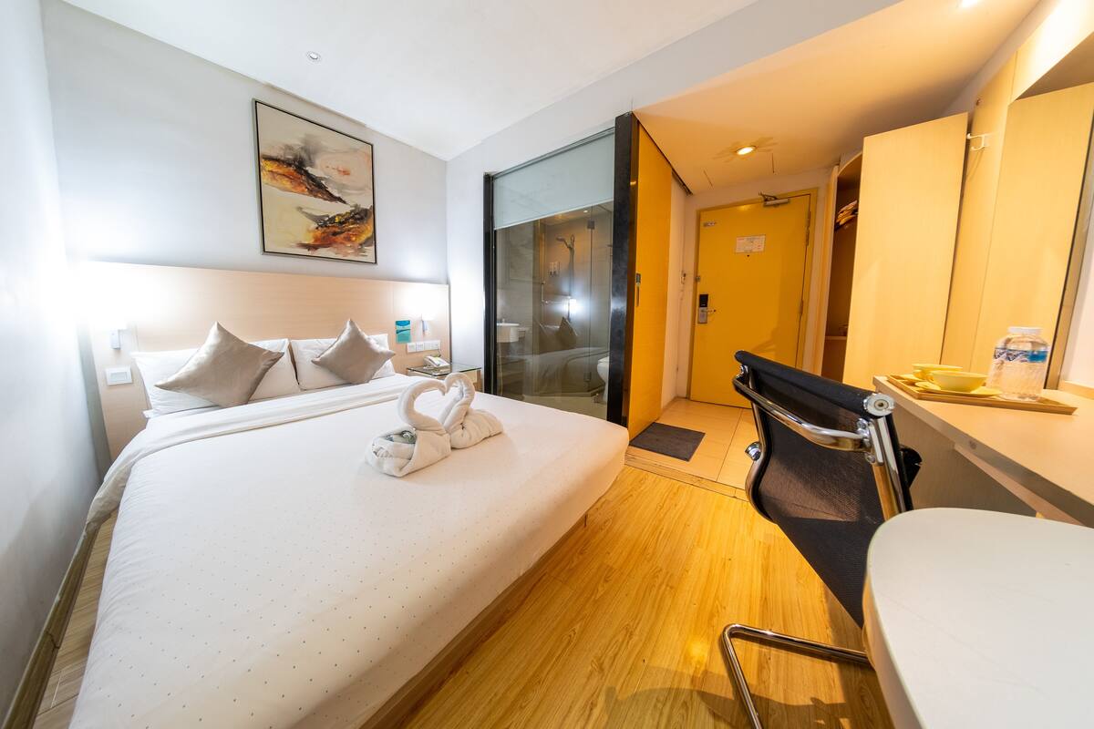A hotel room with a bed, a desk, a chair, a bathroom with a glass door, and a painting on the wall.