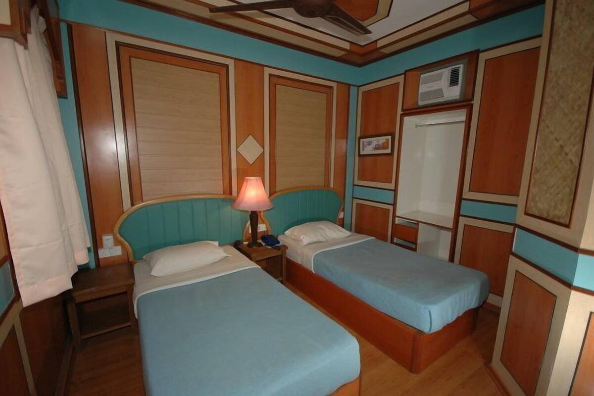 A room with two single beds, a nightstand with a lamp, a wardrobe, and an air conditioning unit.