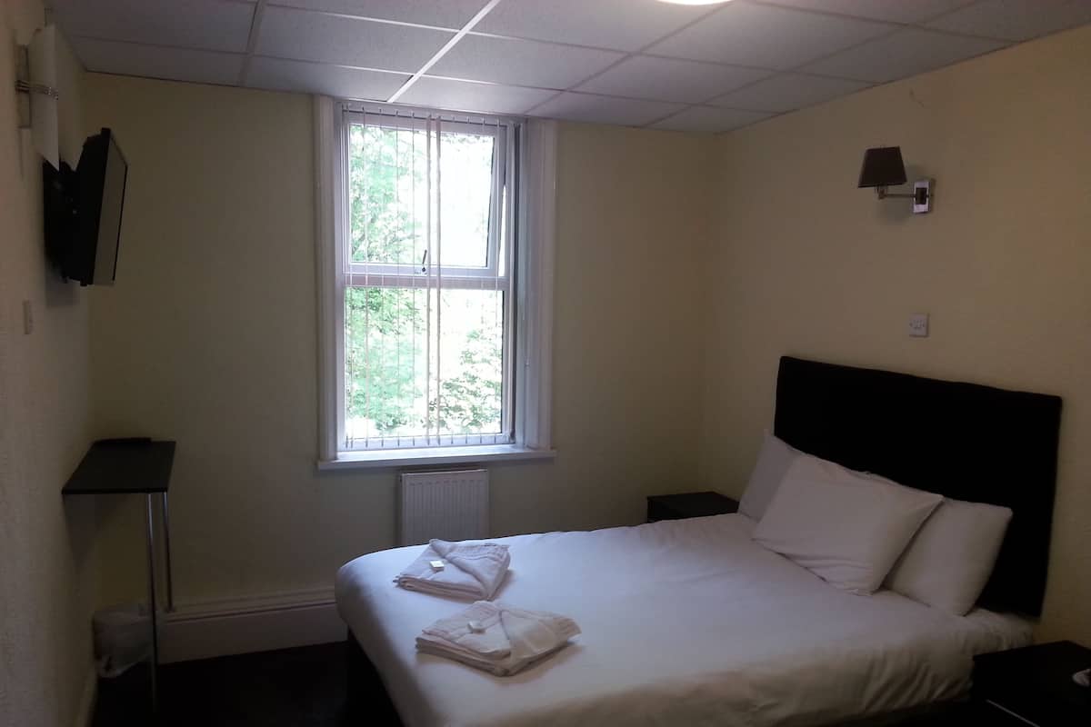 Quarto de hotel com cama, janela, mesa de cabeceira e luminária fixada na parede.