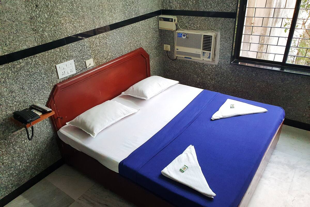 A hotel room with a bed, pillows, a bedside phone, an air conditioning unit, and a window with bars.