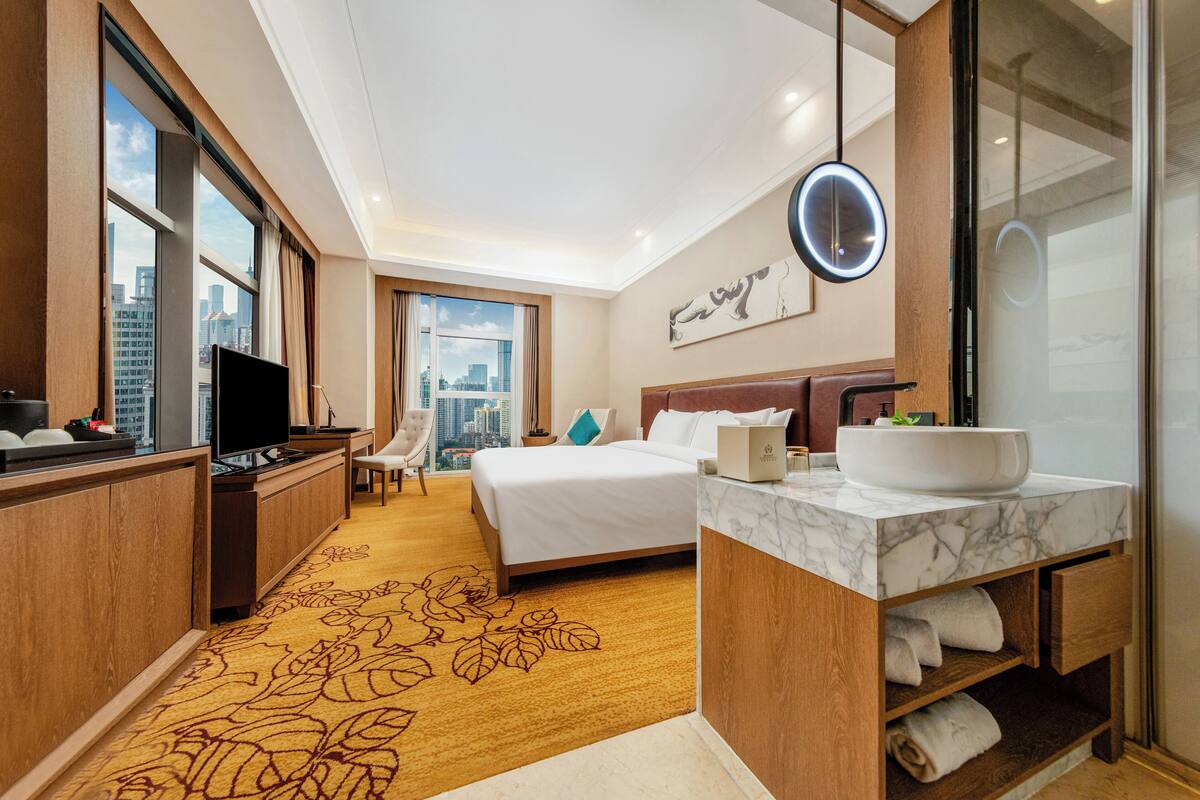Una habitación de hotel con una cama, un escritorio con televisor, un baño con lavamanos amplio y vistas a la ciudad a través de las ventanas.