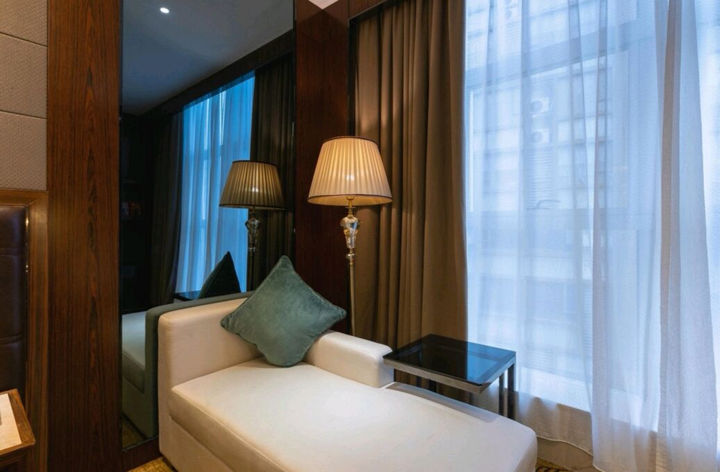 A hotel room with a sofa, a small table, a lamp, and a window with curtains.