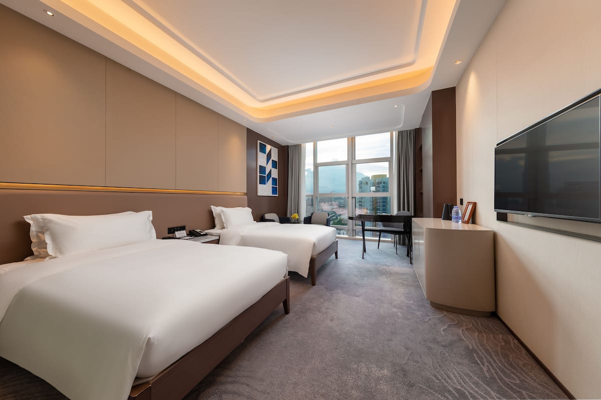 Habitación de hotel con dos camas, un escritorio, un televisor y un ventanal con vistas a la ciudad.