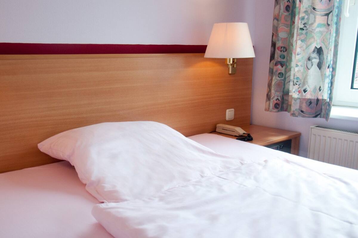 Habitación de hotel con cabecera de madera, lámpara de noche, teléfono y ventana con cortinas.