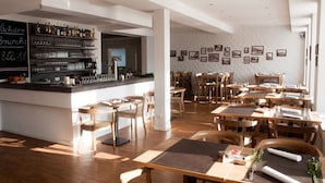 A restaurant interior with wooden tables and chairs, a bar area with shelves of bottles, and framed pictures on the walls.