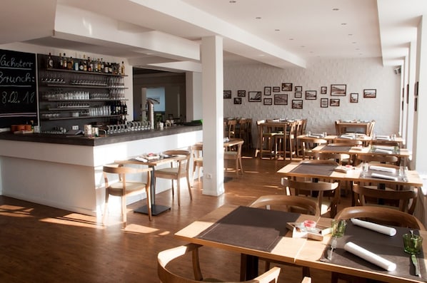 Un interior de restaurante con mesas y sillas de madera, una zona de bar con estantes llenos de botellas y cuadros en las paredes.