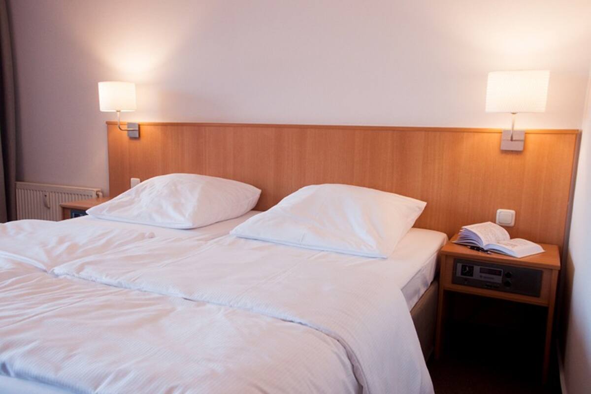 Una habitación de hotel con cama doble, dos lámparas de noche, una mesita de noche con un libro y una radio, y un radiador.