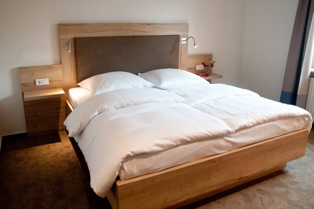 Una cama grande con cabecera de madera, una mesita de noche con lámpara y una mesita con un jarrón de flores.