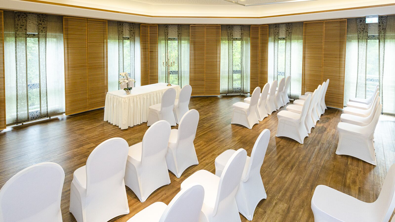 A conference room with rows of white chairs and a table, large windows with blinds, and a wooden floor.