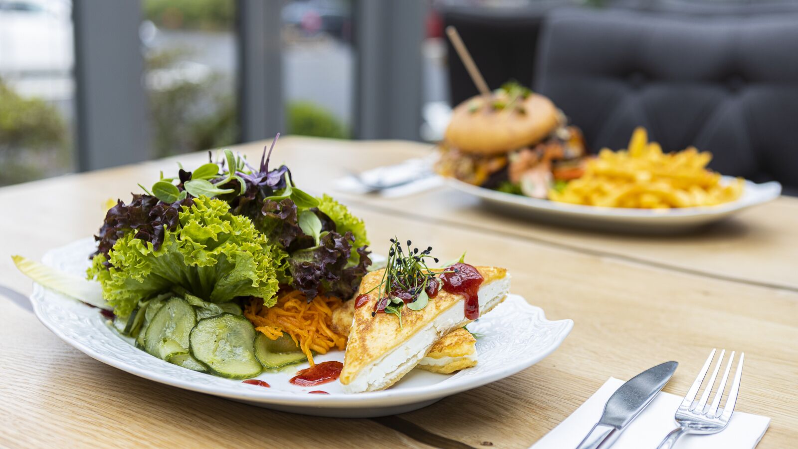 Una mesa de comedor con platos de comida, incluyendo una ensalada y un sándwich, y un tenedor y un cuchillo a la derecha.