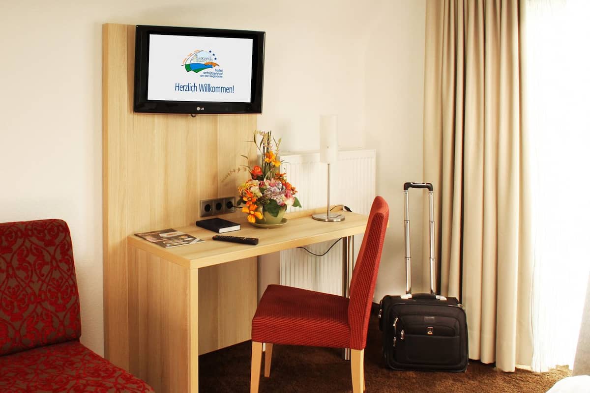 Una habitación de hotel con un escritorio de madera, una silla roja, una maleta negra, un televisor mostrando un mensaje de bienvenida y un arreglo floral.