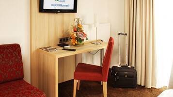 Una habitación de hotel con un escritorio de madera, una silla roja, una maleta negra, un televisor mostrando un mensaje de bienvenida y un arreglo floral.