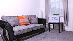 Ein graues Sofa mit einem orange-grauen Kissen, ein kleiner schwarzer Nachttisch mit einem Schild und ein Fenster mit durchsichtigen Vorhängen.